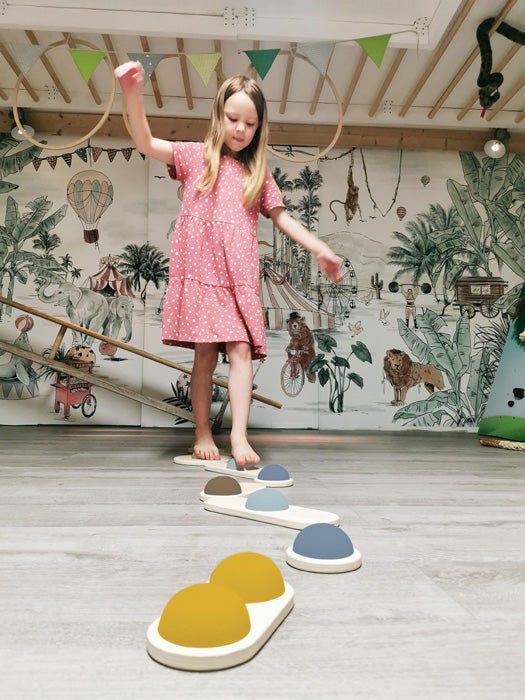 Ein junges Mädchen im rosa Kleid balanciert auf Balanciersteinen in einem Spielzimmer mit einer Wandmalerei zum Thema Dschungel und verbessert so spielerisch ihren Gleichgewichtssinn.