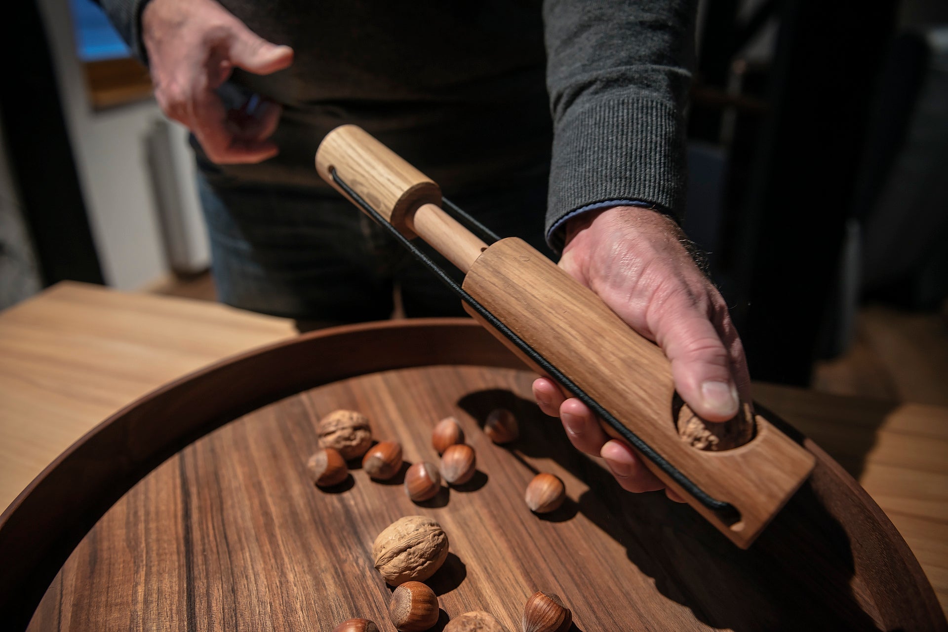 Mit dem Nussknacker Nuss Kracher aus Eichenholz knackt man eine Walnuss über einem Tablett voller Nüsse.