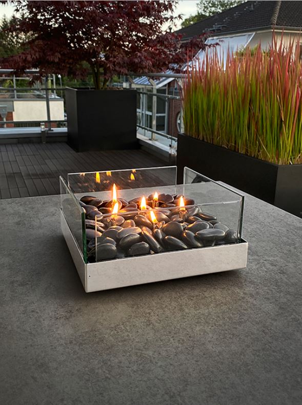 Ein Design-Tischkamin aus Edelstahl mit schwarzen Steinen und Glasscheiben brennt auf einem Terrassentisch nahe hoher Pflanzen und eines roten Baums – befeuert mit Paraffinöl für stimmungsvolles Ambiente.