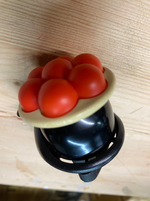 Die Schwarzwald Bollenhut Fahrradklingel, eine schwarze Fahrradklingel aus Kunststoff mit roten, blasenartigen Akzenten und beigem Rand, sitzt auf einer Holzoberfläche und bringt einen authentischen Hauch von Schwarzwaldflair.