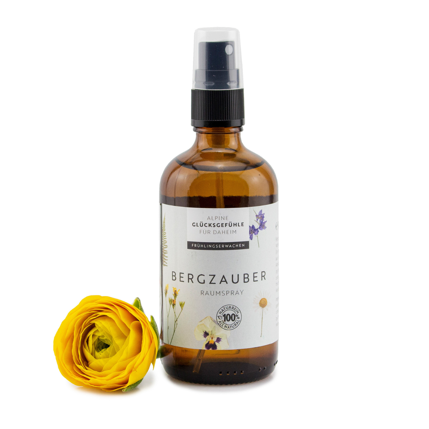 Eine braune Glassprühflasche mit der Aufschrift „Raumspray Bergzauber“ steht neben einer gelben Ranunkelnblüte, die ätherische Öle für eine angenehme Duftneutralisierung im Raum enthält.