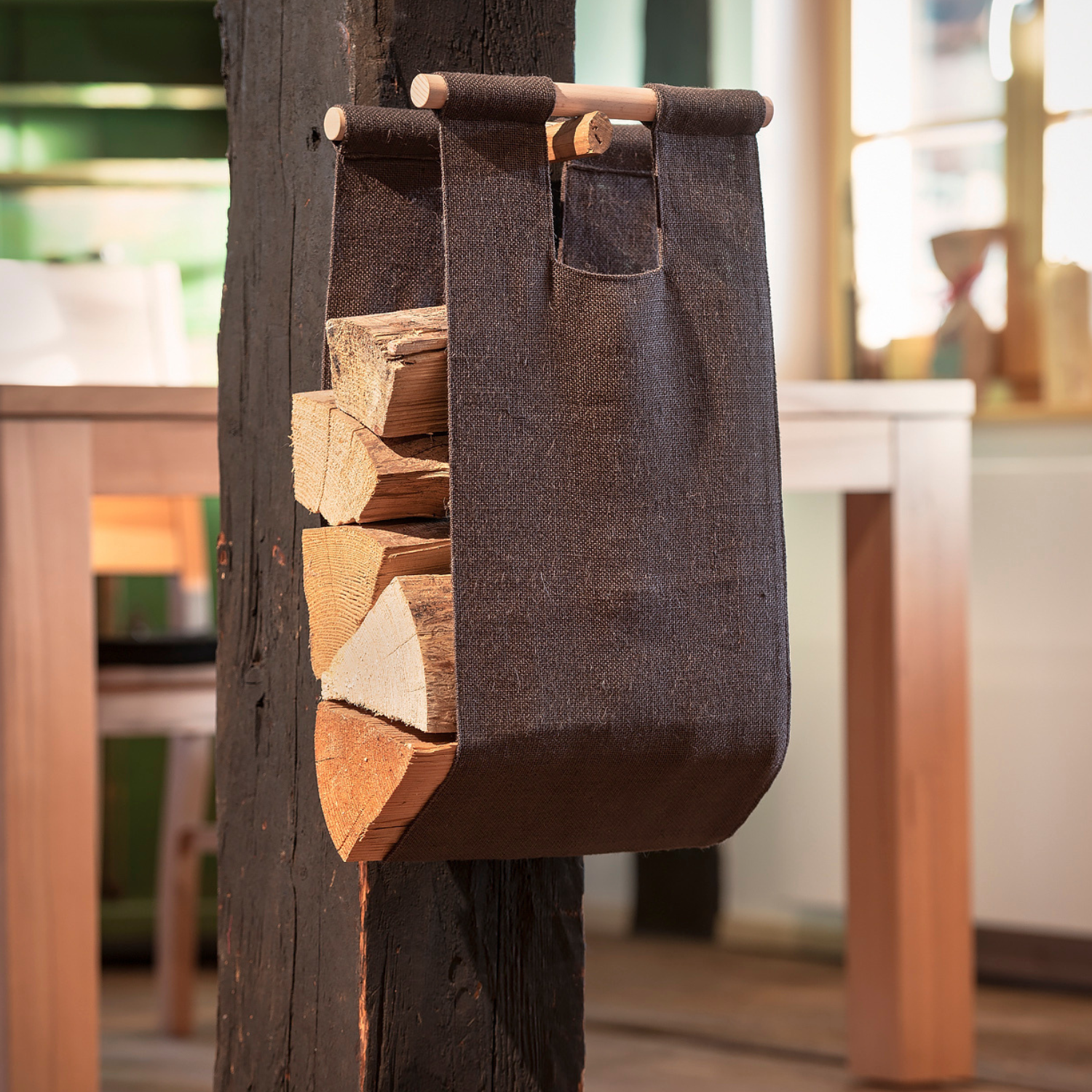 Die Kaminholztrage Feuerzauber aus Jutestoff in Schwarz mit eleganten Buchenholzgriffen hängt an einem dunklen Holzpfosten und hält ordentlich gespaltenes Kaminholz.