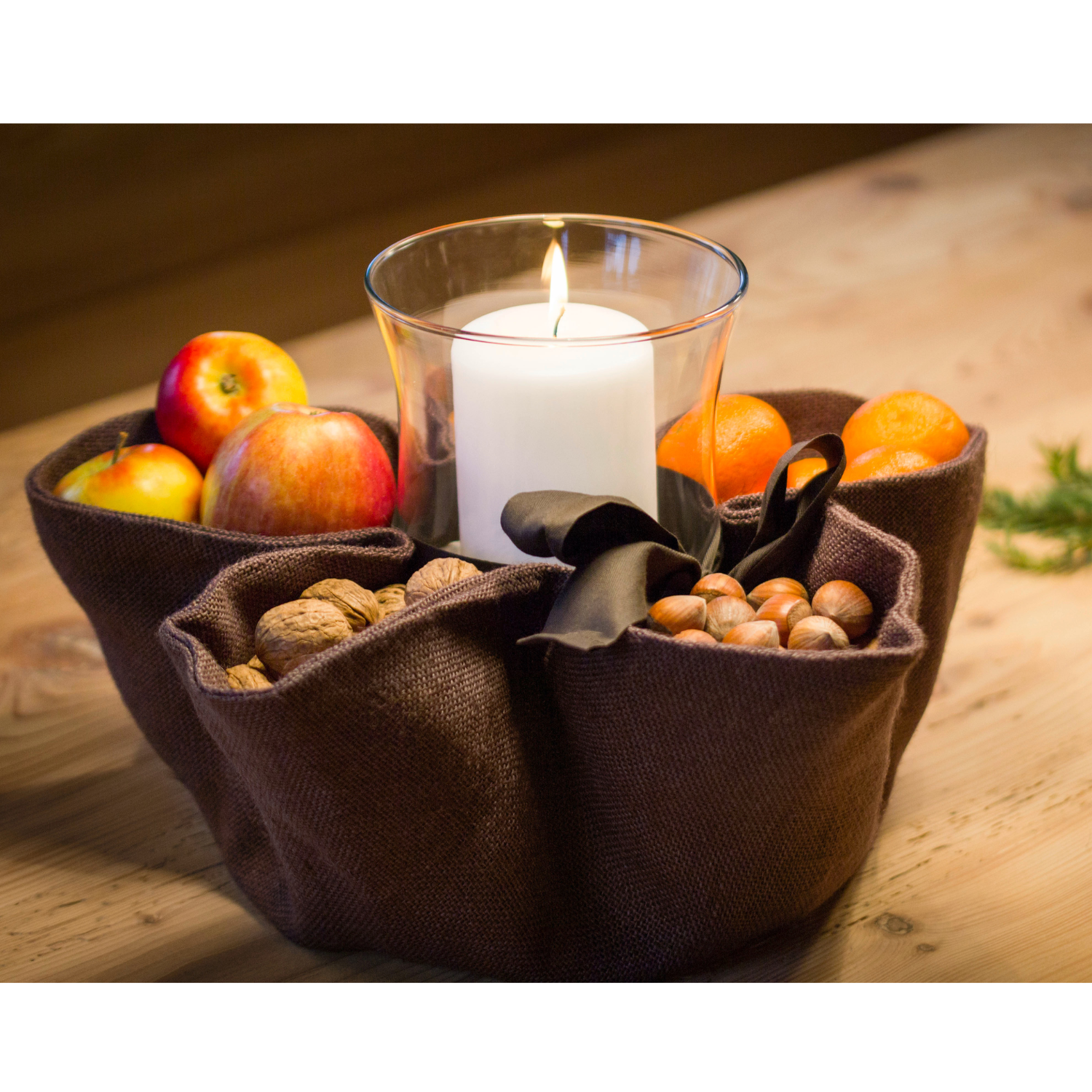 Ein brauner Nusssack mit Äpfeln, Orangen, Nüssen und einer brennenden Kerze steht auf einem Holztisch – ein elegantes Einrichtungsaccessoire für jedes Zuhause.