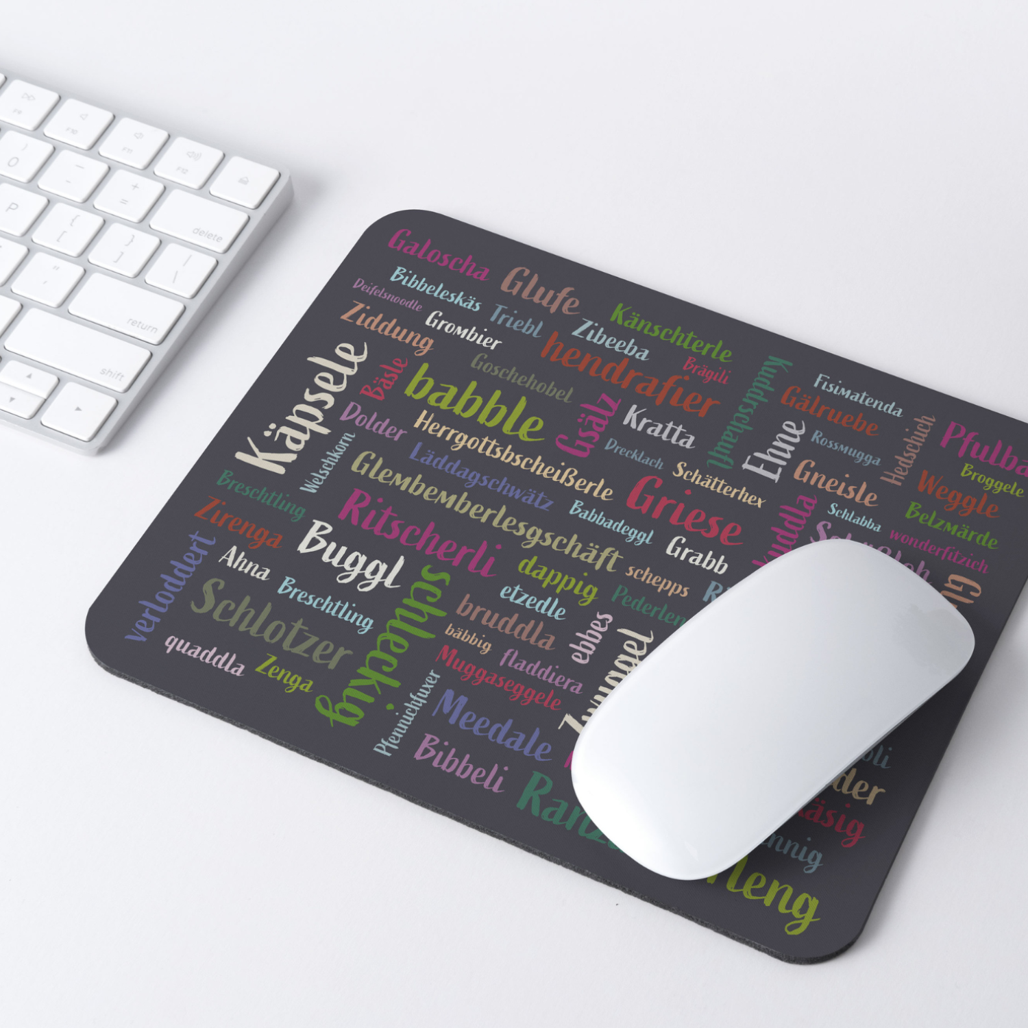 Eine weiße Maus und das Mundart-Mousepad mit bunten deutschen Wörtern im Mundart-Design liegen neben einer kleinen Topfpflanze auf einem weißen Schreibtisch, sorgen für Flair und schützen die Oberfläche.