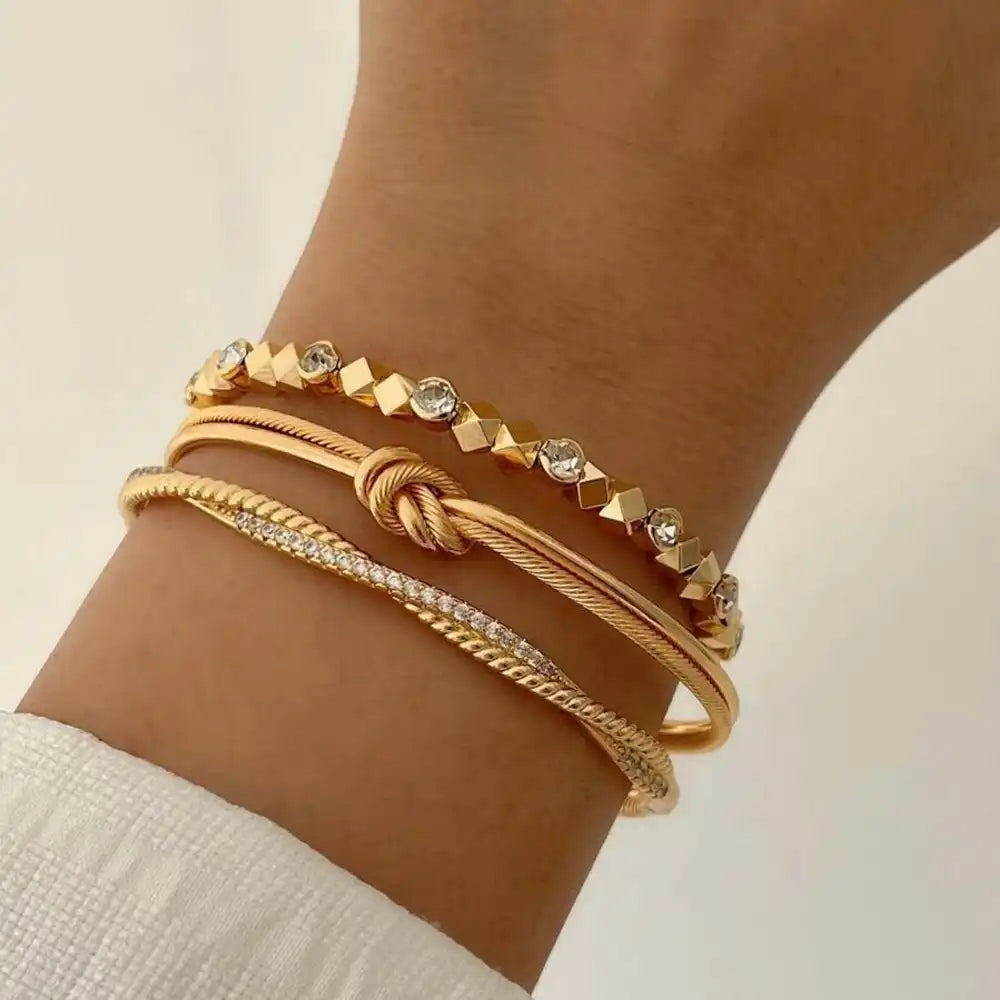 Ein Handgelenk mit drei goldenen Armbändern, eines davon mit Kristallen, vor einem hellen Hintergrund.