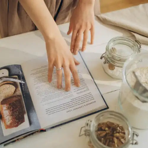 Hände, die ein Kochbuch lesen, neben Gläsern mit Mehl und Nüssen auf einem Küchentisch.