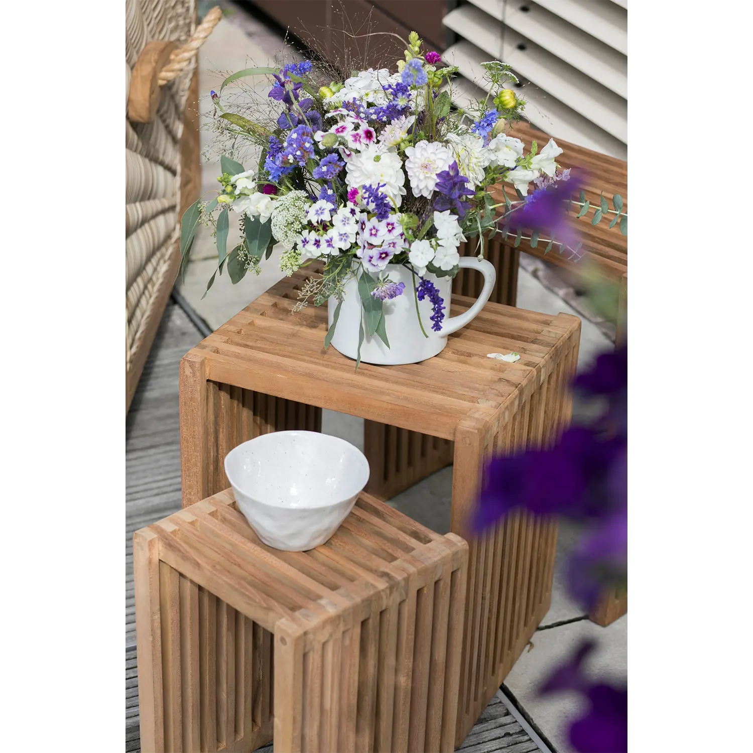 Ein weißer Keramikkrug mit lila-weißen Blumen ziert das Beistelltische 3er-Set, darunter steht eine weiße Schale. Dieses elegante Arrangement ergänzt das Outdoor-Ambiente, umgeben von Flechtmöbeln und Lamellenjalousien.