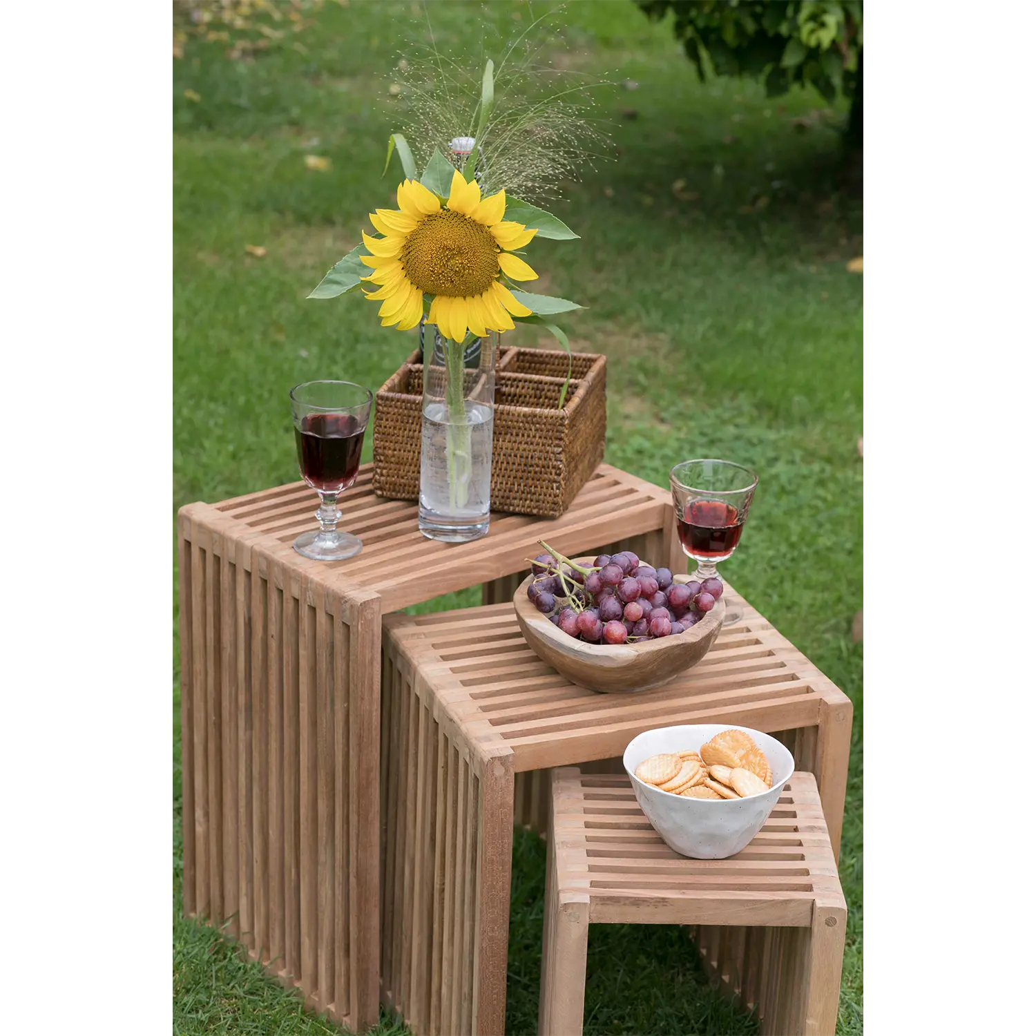 Eine Szene im Freien zeigt ein gestapeltes 3er-Set Beistelltische, bestehend aus einer Vase mit einer Sonnenblume, zwei Rotweingläsern, einer Schale mit Trauben, Crackern und einer Weidenbox. Diese Tische aus hochwertigem Teakholz ruhen elegant auf dem üppigen grünen Gras.
