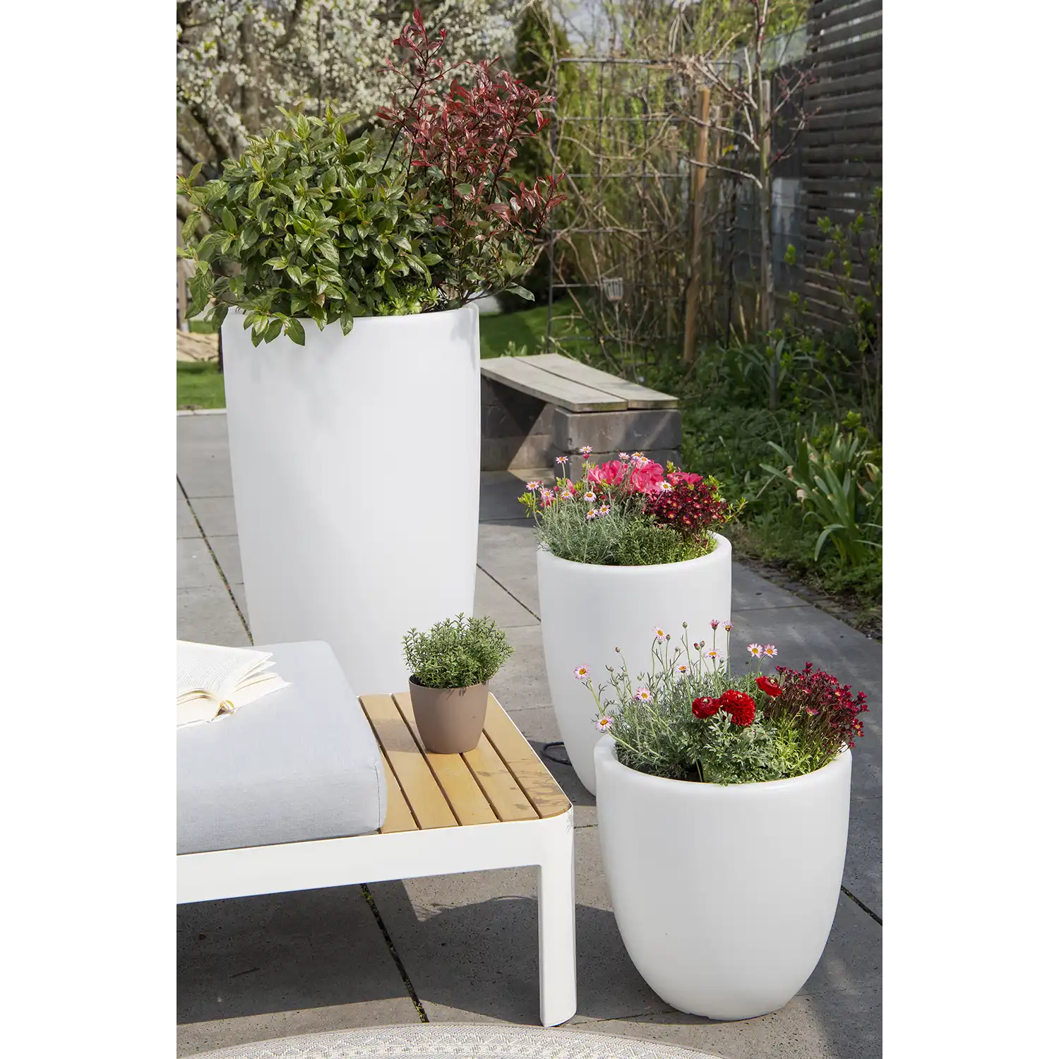 Drei weiße, beleuchtete Blumentöpfe in unterschiedlichen Höhen, gefüllt mit bunten Blumen und Grünpflanzen, sind auf einer Terrasse neben einer Bank mit Kissen und offenem Buch angeordnet und schaffen so einen stilvollen, wetterfesten Sitzbereich im Freien.