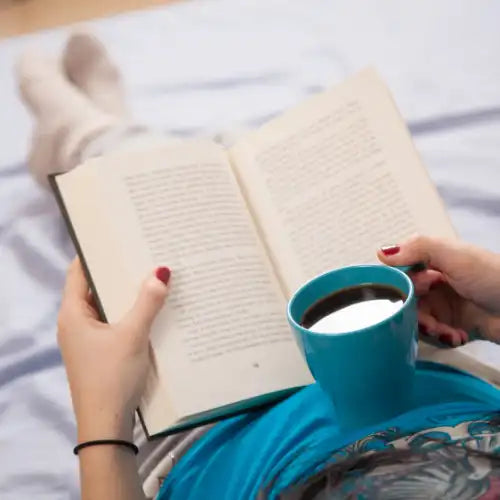Eine Person liest ein Buch und hält einen blauen Kaffeebecher in der Hand, während sie sich auf einem Bett ausruht.