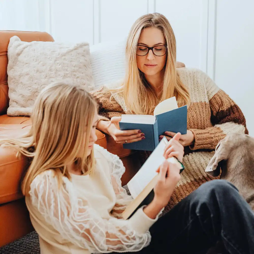 Zwei Personen sitzen zusammen und lesen Bücher auf dem Boden neben einem braunen Sofa.