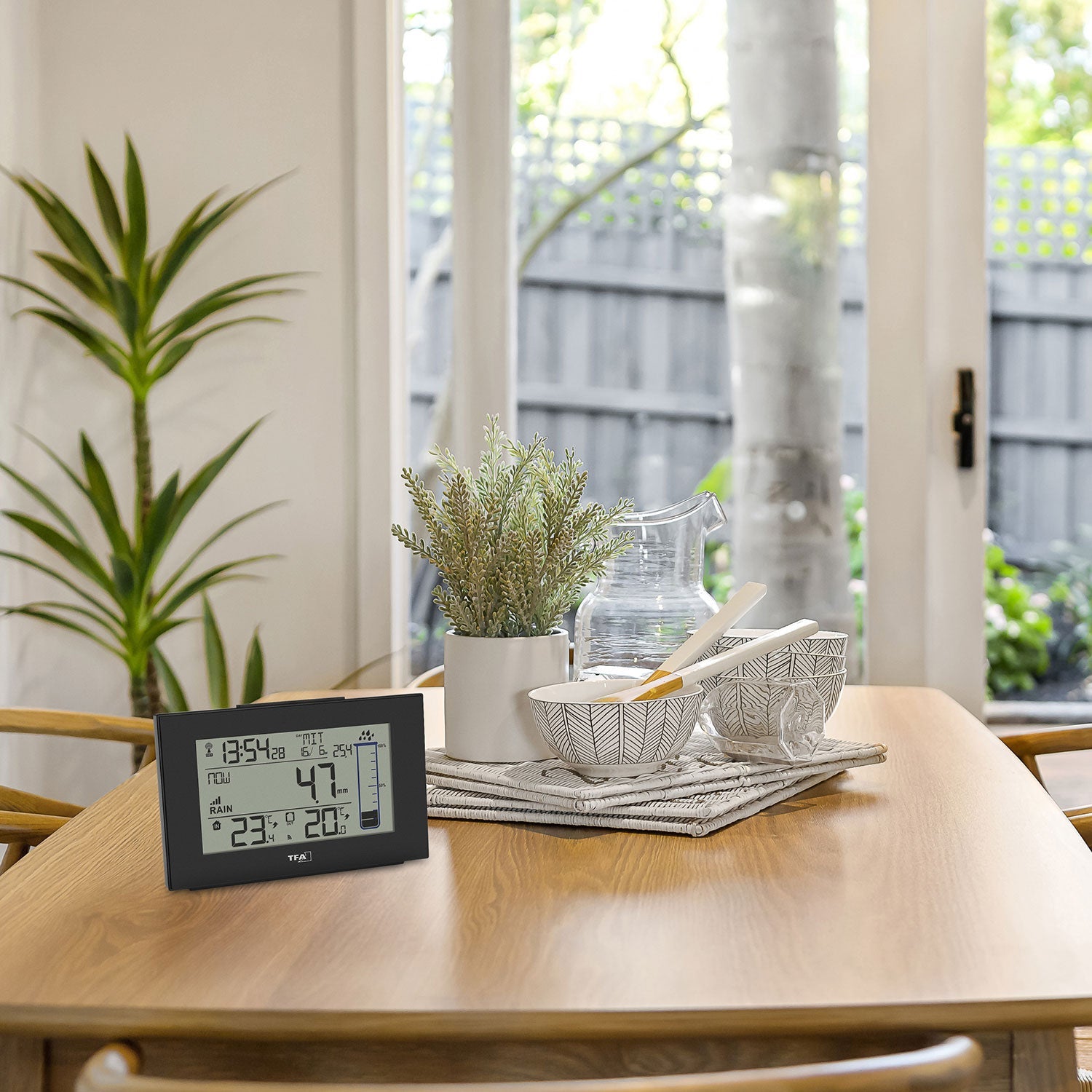 Auf einem Holztisch steht ein digitaler Funk-Regenmesser, der Temperatur und Luftfeuchtigkeit anzeigt. Flankiert wird er von zwei gemusterten Schalen, einem Wasserkrug aus Glas, einem gefalteten Tuch und einer Topfpflanze. Durch das Fenster blickt man auf einen ruhigen Baum und einen Zaun.