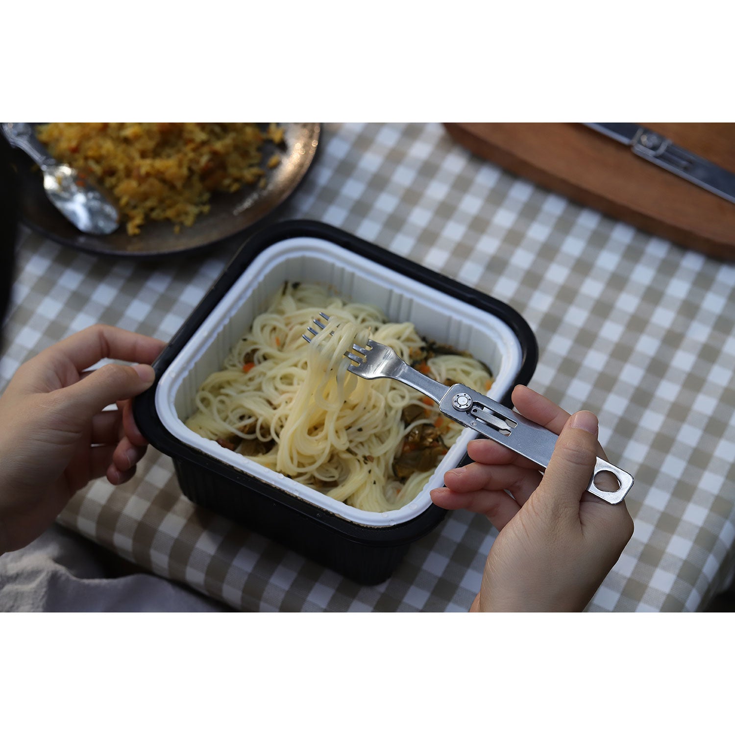 Jemand hält ein elegantes Edelstahl-Multitool mit Gabel in der Hand und genießt Pasta aus einem Behälter. Der schwarz-weiße Behälter steht auf einer karierten Tischdecke, im Hintergrund ein weiterer Teller mit Essen und einer zusätzlichen Gabel. Produktname: Faltbares Besteck / Camping Besteck.