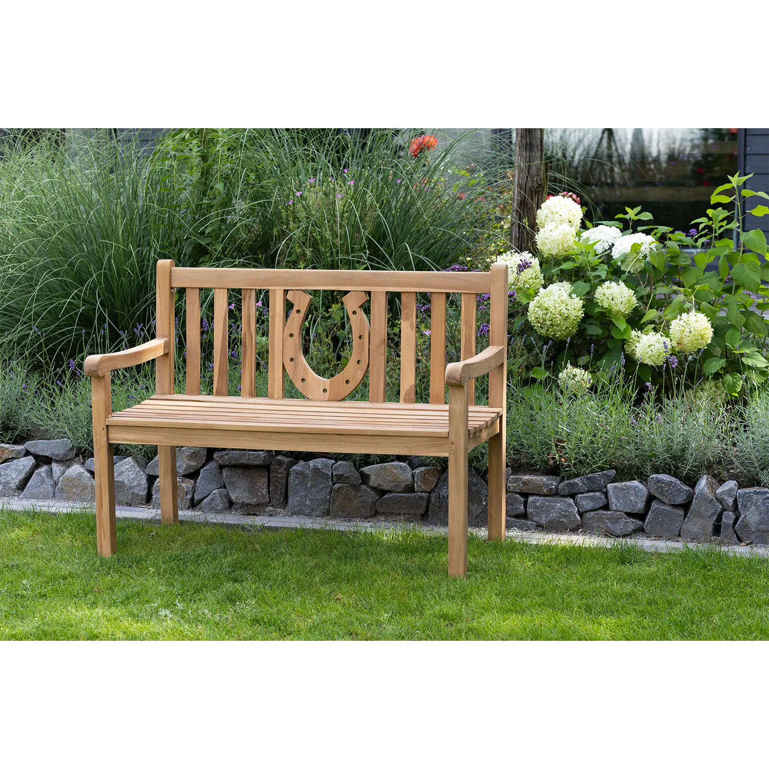 Die Teak-Gartenbank Hufeisen liegt elegant auf einer Rasenfläche, eingerahmt von dekorativen Steinkanten, üppigem Grün und weißen Blumen, und bietet so einen ruhigen Rückzugsort im Garten.