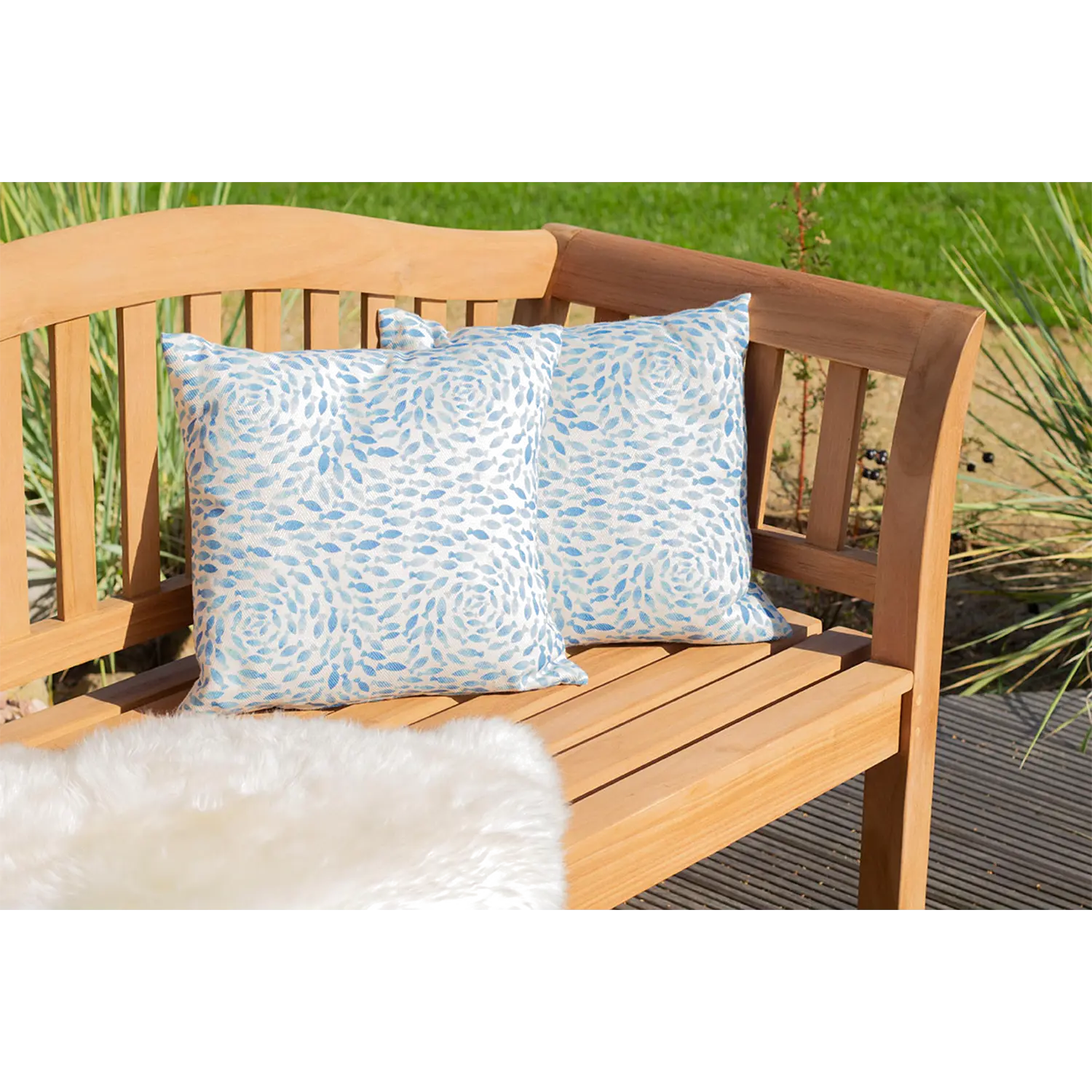 Die Teak-Gartenbank Wave, geschmückt mit blauen und weißen Kissen, ist mit einer flauschigen weißen Decke bedeckt und sorgt so für eine gemütliche Sitzgelegenheit. Sie ruht auf einem Deck inmitten üppigen Grases und Pflanzen unter der strahlenden Sonne.