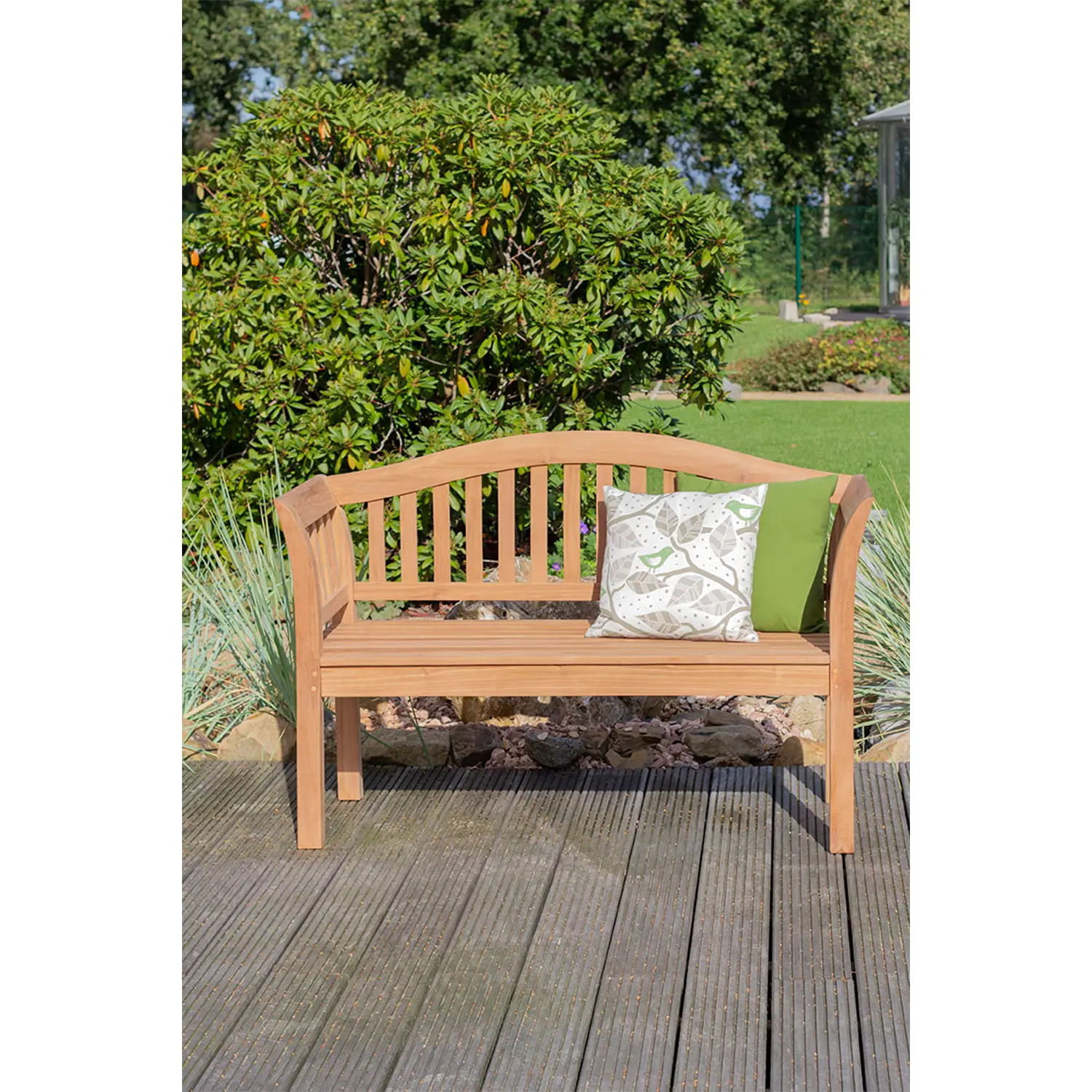 Eine Teak-Gartenbank Wave mit einem grünen und einem gemusterten Dekokissen steht auf einem Holzdeck, umgeben von Büschen, Gras und Bäumen und schafft so einen ruhigen Sitzbereich im Freien.