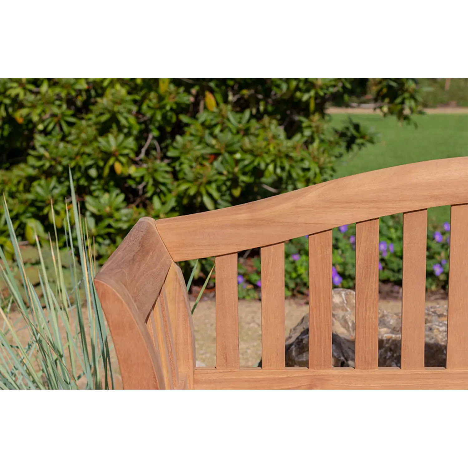 Eine Nahaufnahme der Rückenlehne der Teak-Gartenbank Wave im Außenbereich zeigt ihr Lattendesign inmitten üppiger grüner Pflanzen und Gräser, mit Rasen und Bäumen als ruhigem Hintergrund. Helles Sonnenlicht hebt die natürliche Maserung dieser eleganten Sitzgelegenheit hervor.