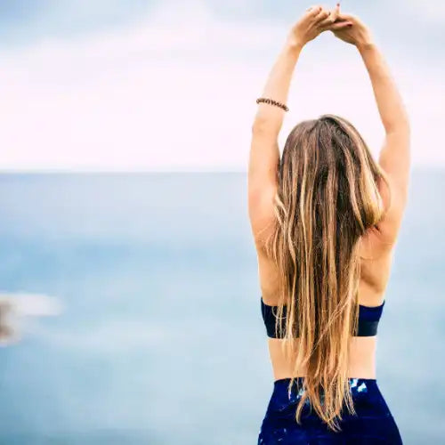 Frau mit langen Haaren, die ihre Arme über den Kopf streckt, am Meer.