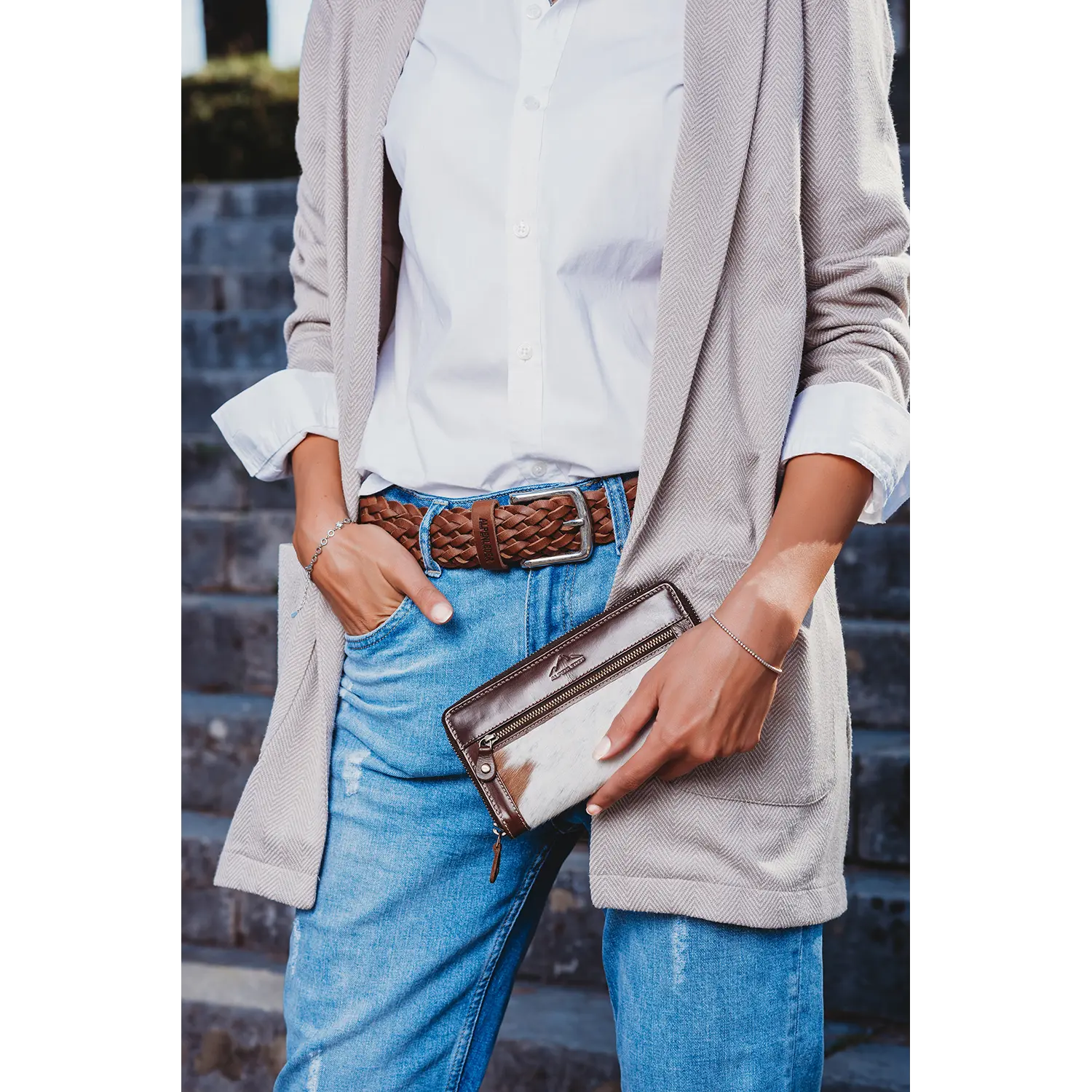 Eine Person in weißem Hemd, beiger Strickjacke und Bluejeans steht auf einer Steintreppe und hält den Damengeldbeutel GSPUSI in der Hand, eine braun-weiße Clutch aus Kuhfell. Eine Hand in der Tasche trägt sie einen braunen geflochtenen Gürtel, der Stil mit RFID-Schutz verbindet.