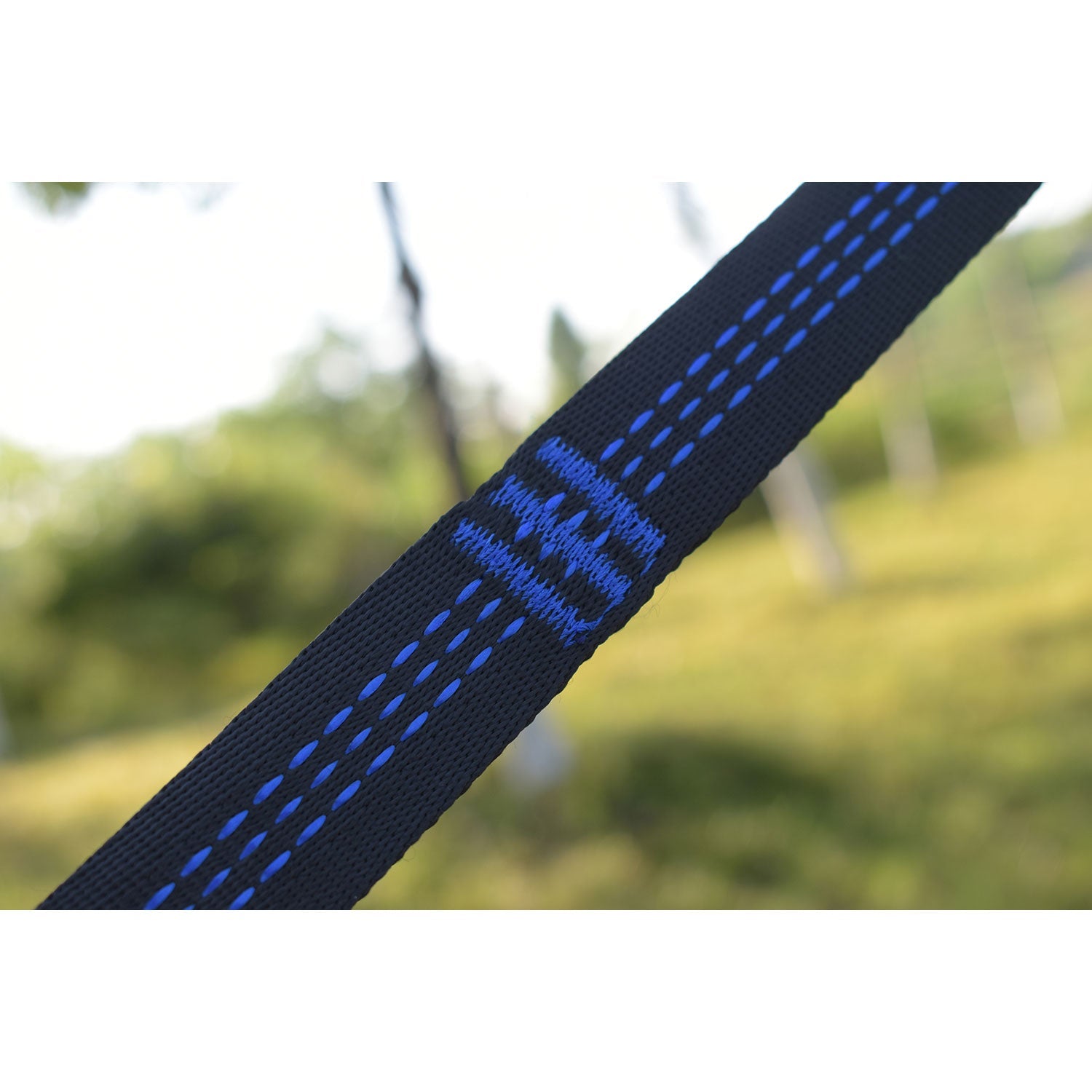 Die Nahaufnahme eines gespannten Gurtes mit blauen Nähten verdeutlicht seine Strapazierfähigkeit. Im unscharfen Hintergrund suggerieren Gras und Bäume einen Park oder eine natürliche Umgebung – perfekt für die Superleichte Hängematte.