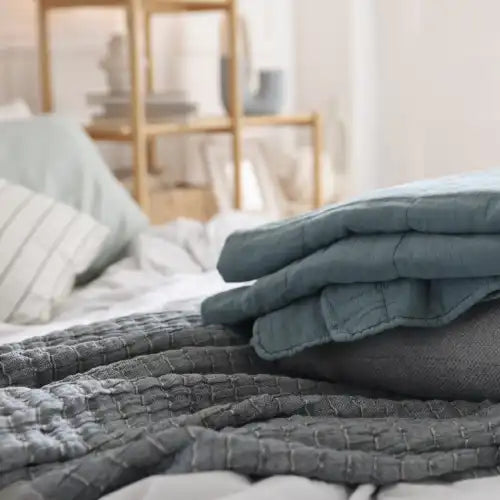 Gefaltete blaue und graue Steppdecken auf einem ordentlich gemachten Bett in einem gemütlichen Schlafzimmer.