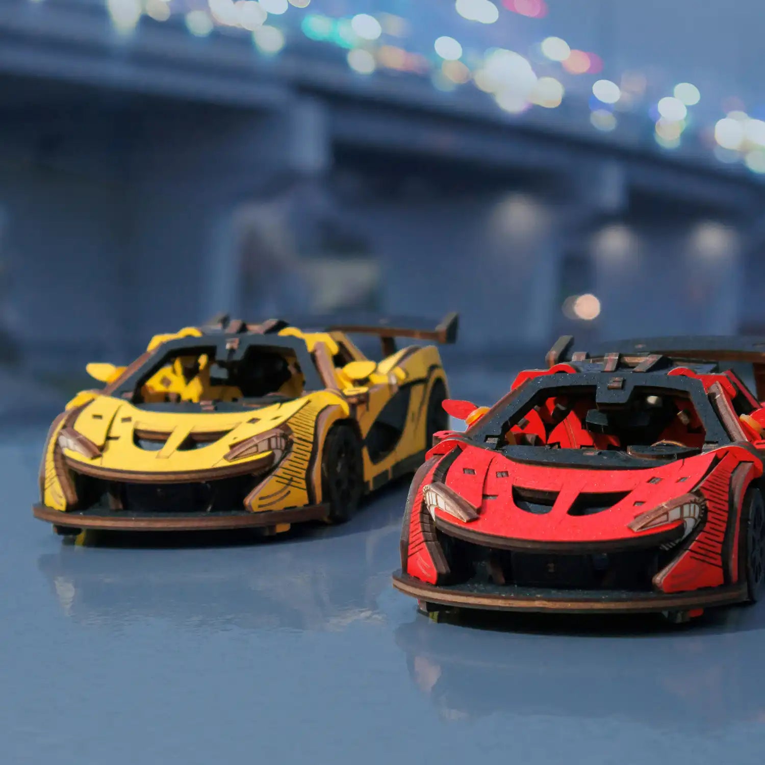Das 3D-Holzpuzzle Rennwagen gelb begeistert Modellbau-Fans mit seiner filigranen Gestaltung, die neben einem roten Pendant auf einer spiegelnden Oberfläche thront. Die Szenerie vor einer beleuchteten Nachtbrückenkulisse ist ideal für Liebhaber detailreicher Rennwagenmodelle.