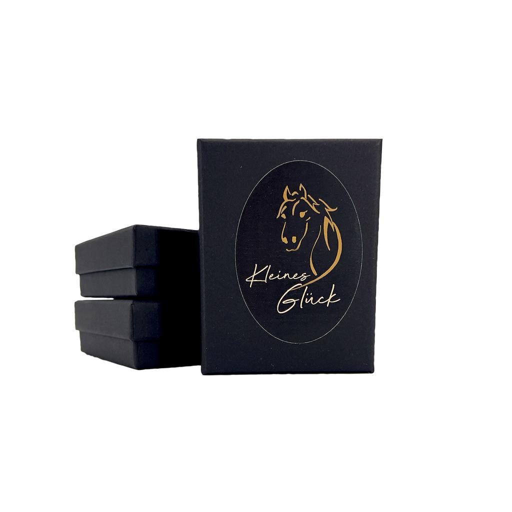 Neben einer aufrecht stehenden Box steht ein Stapel aus drei schwarzen Kisten, auf denen jeweils ein goldenes Pferdekopf-Logo und die Aufschrift „Kleines Glück“ in Kursivschrift zu sehen sind. Dies verweist auf die Verbindung zum Hufnagel Schmuck mit Lavastein grün und Engelsflügel, der das zeitlose Glückssymbol eines Hufeisens verkörpert.