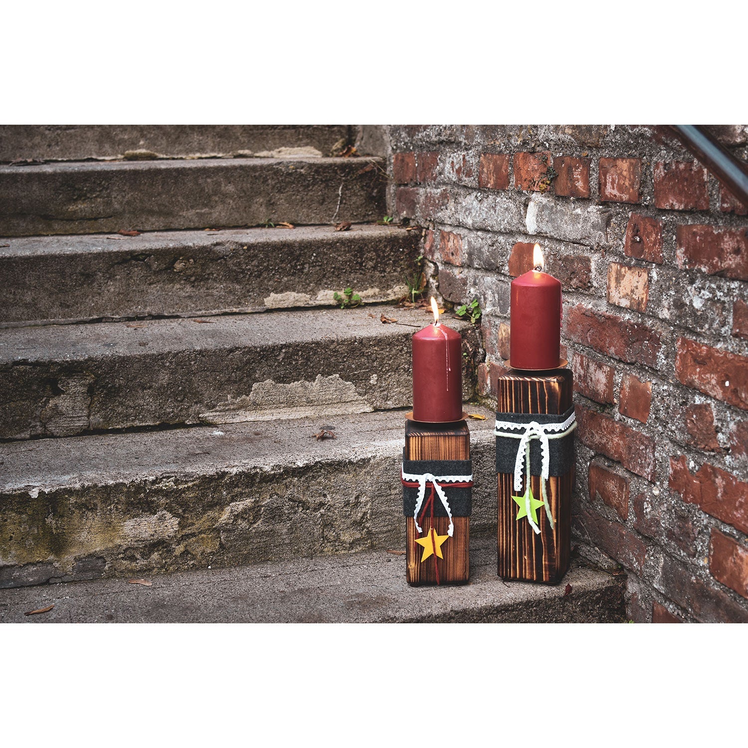 Zwei rote Stumpenkerzen, beleuchtet auf Kerzenständern aus Holz – groß mit Sternendekor, beleuchten die Stufen neben einer roten Backsteinmauer. Der verwitterte Stein trägt zum rustikalen Ambiente bei.