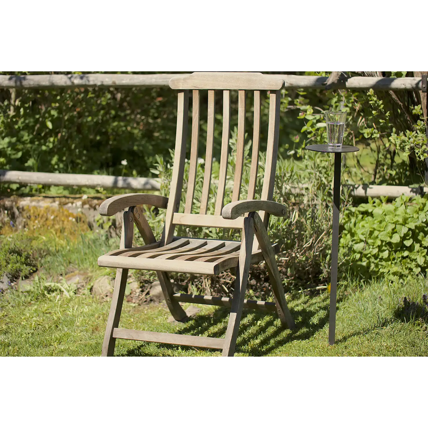 Ein hölzerner Gartenstuhl steht auf einer Wiese in einem Garten, neben einem kleinen MONO-Gartentischchen mit einem Glas Wasser. Im Hintergrund sind grüne Pflanzen und ein Holzzaun mit rostiger Patina zu sehen.