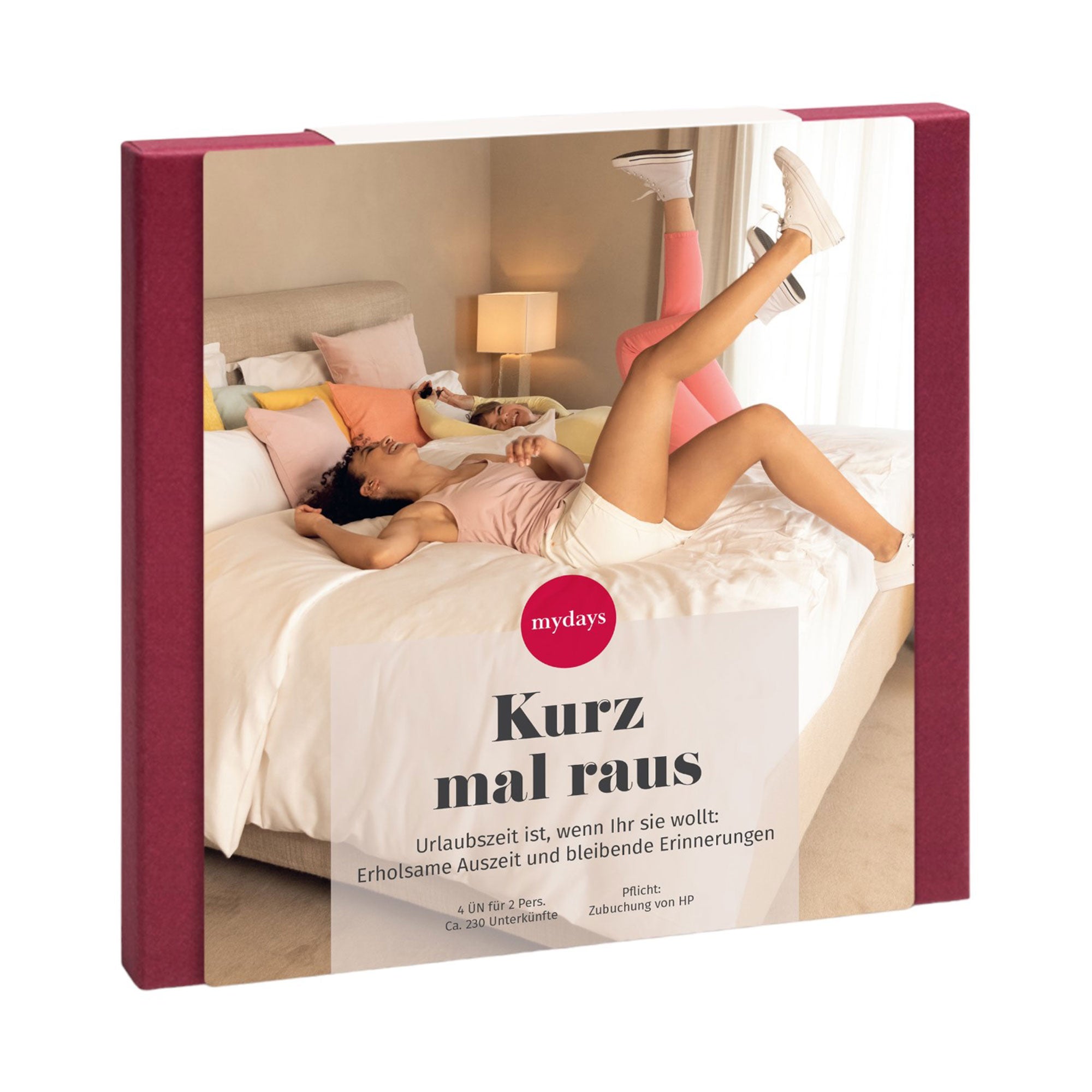 Eine Frau lümmelt verspielt auf einem Bett, umgeben von bunten Kissen in einem gemütlichen Zimmer. Die „Erlebnis-Box Kurz mal raus“ bietet die perfekte Idee für eine erholsame Auszeit.