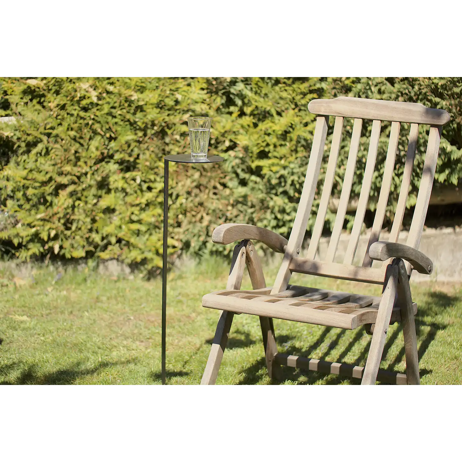 Ein Gartenstuhl aus Holz steht auf dem Gras neben dem MONO-Gartentischchen mit Rostpatina und hält ein Glas Wasser, im Hintergrund sind an einem sonnigen Tag grüne Büsche zu sehen.