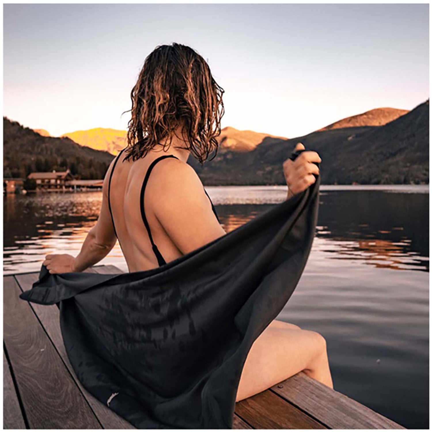 Eine Person mit lockigem Haar sitzt auf einem Holzsteg an einem ruhigen See und hält ein ultraleichtes Nanodry Handtuch in der Hand. Sie blickt auf die Berge, die im warmen Licht des Sonnenuntergangs getaucht sind, während sich die ruhige Szenerie auf der Wasseroberfläche spiegelt.