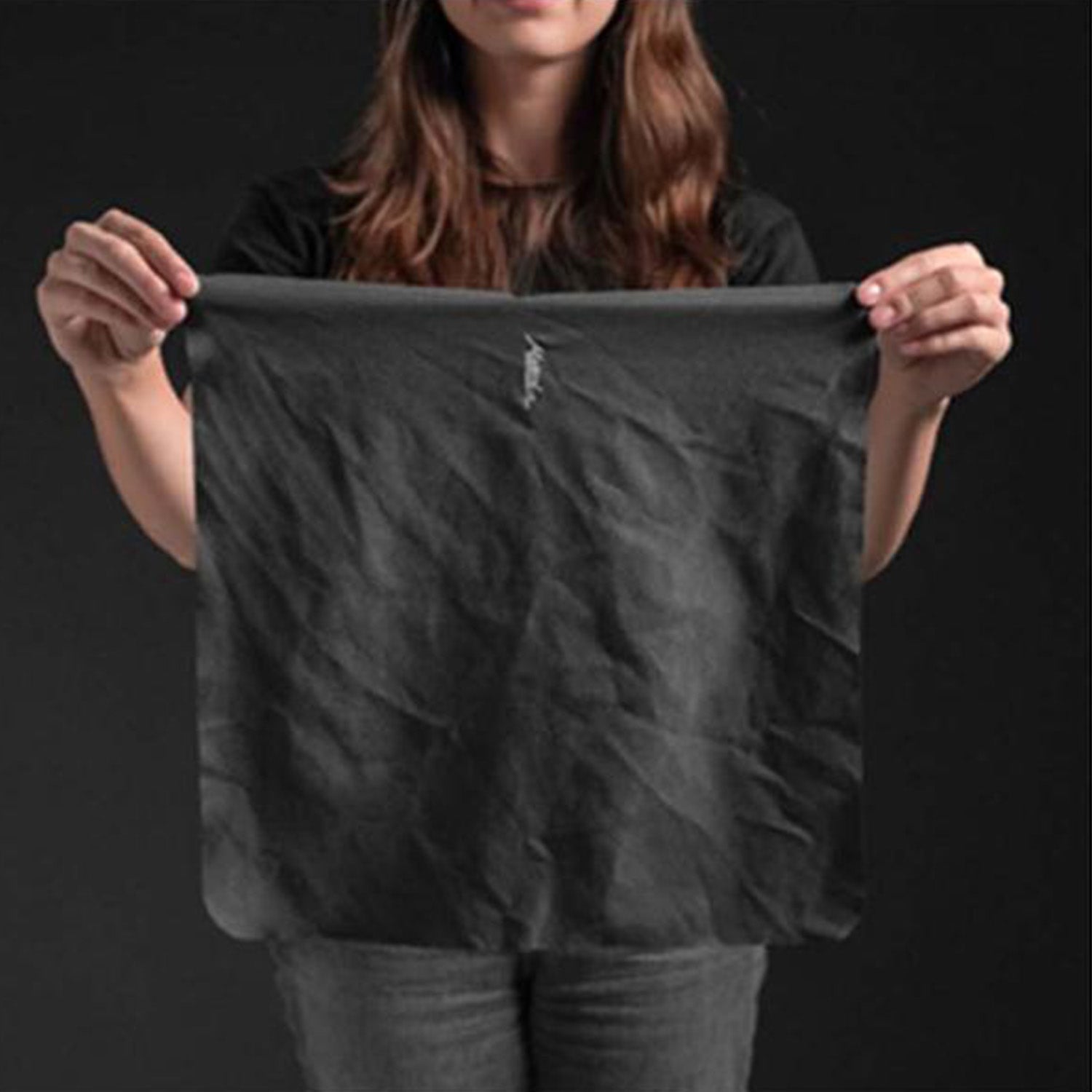 Jemand hält ein großes, dünnes Matador Nanodry Reisehandtuch in beiden Händen vor dunklem Hintergrund. Der zerknitterte Stoff unterstreicht das schlichte Design. In einem schwarzen Oberteil und einer grauen Hose präsentiert er dieses unverzichtbare Outdoor-Equipment perfekt.