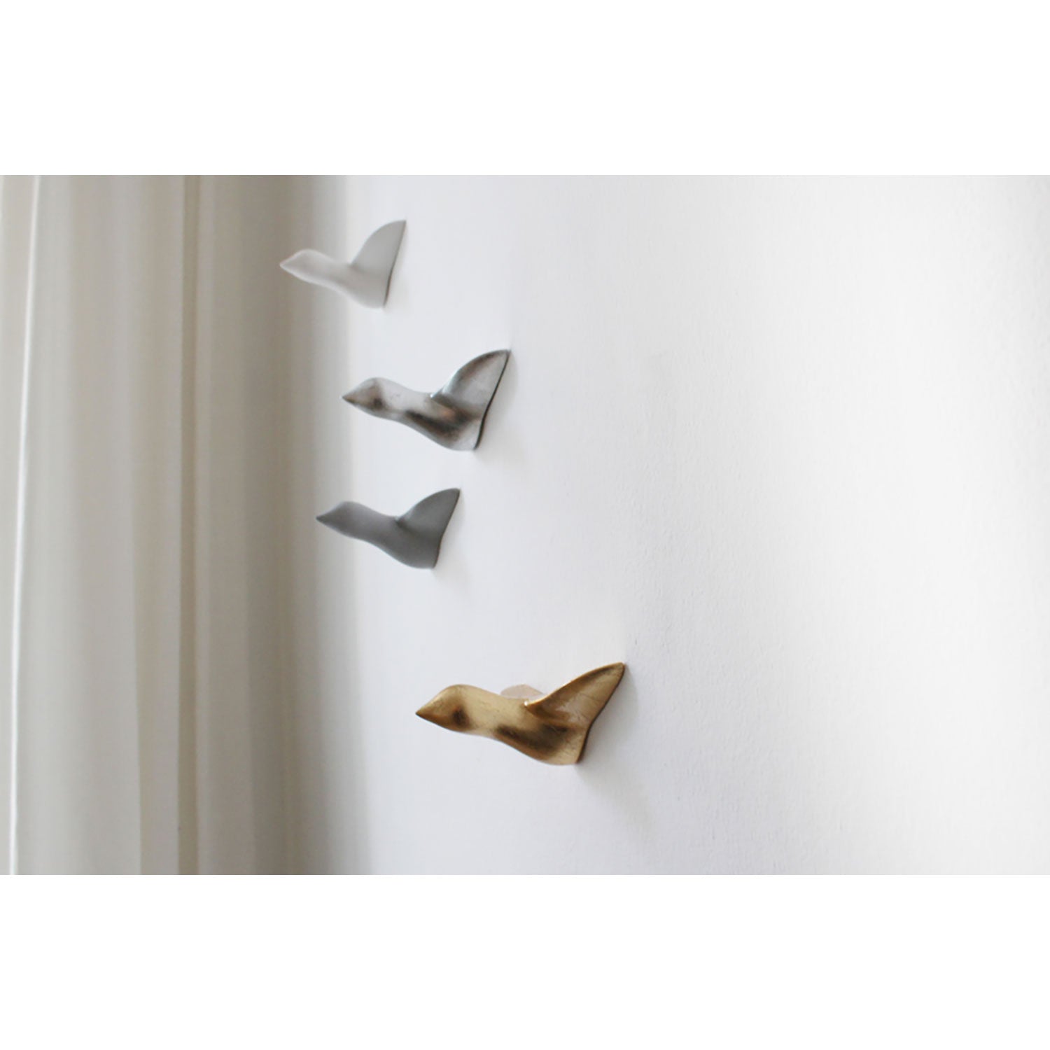 Das Set „Wandskulptur – gemischter Vogelschwarm“ (4 Stk., weiß, gold, grau und silber) verleiht der weißen Wand eine elegante Gestaltung, wobei ein weißer Vorhang teilweise nach links drapiert ist und die künstlerische Handarbeit dieses charmanten Arrangements unterstreicht.