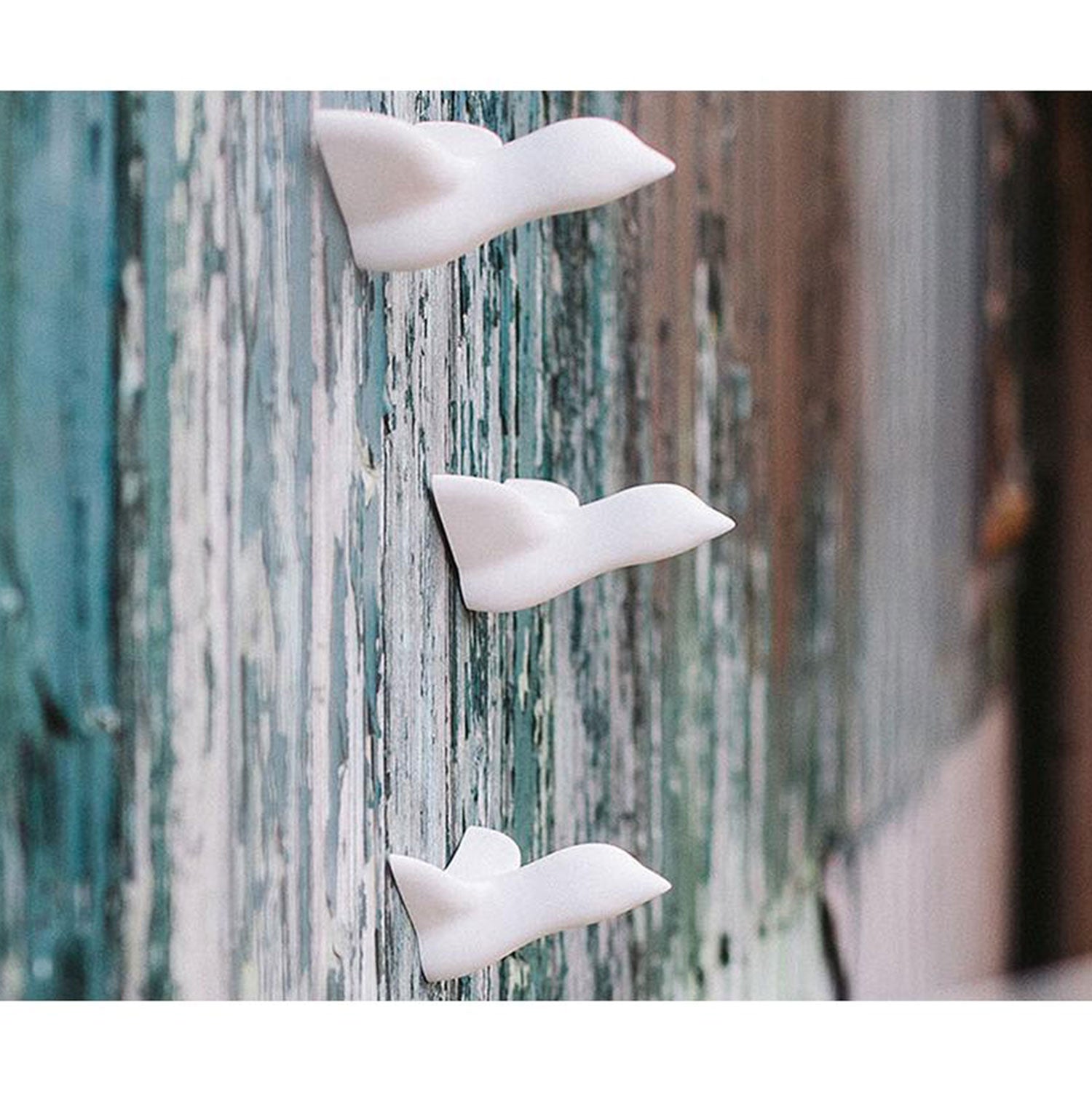 Drei Wandskulpturen vom Typ „Mini Vogel“ sind vertikal auf einer verwitterten blaugrünen und braunen Holzoberfläche montiert und erzeugen den Eindruck von Vögeln im Flug. So entsteht eine einzigartige Wanddekoration, die sowohl dekorativ als auch funktional ist.