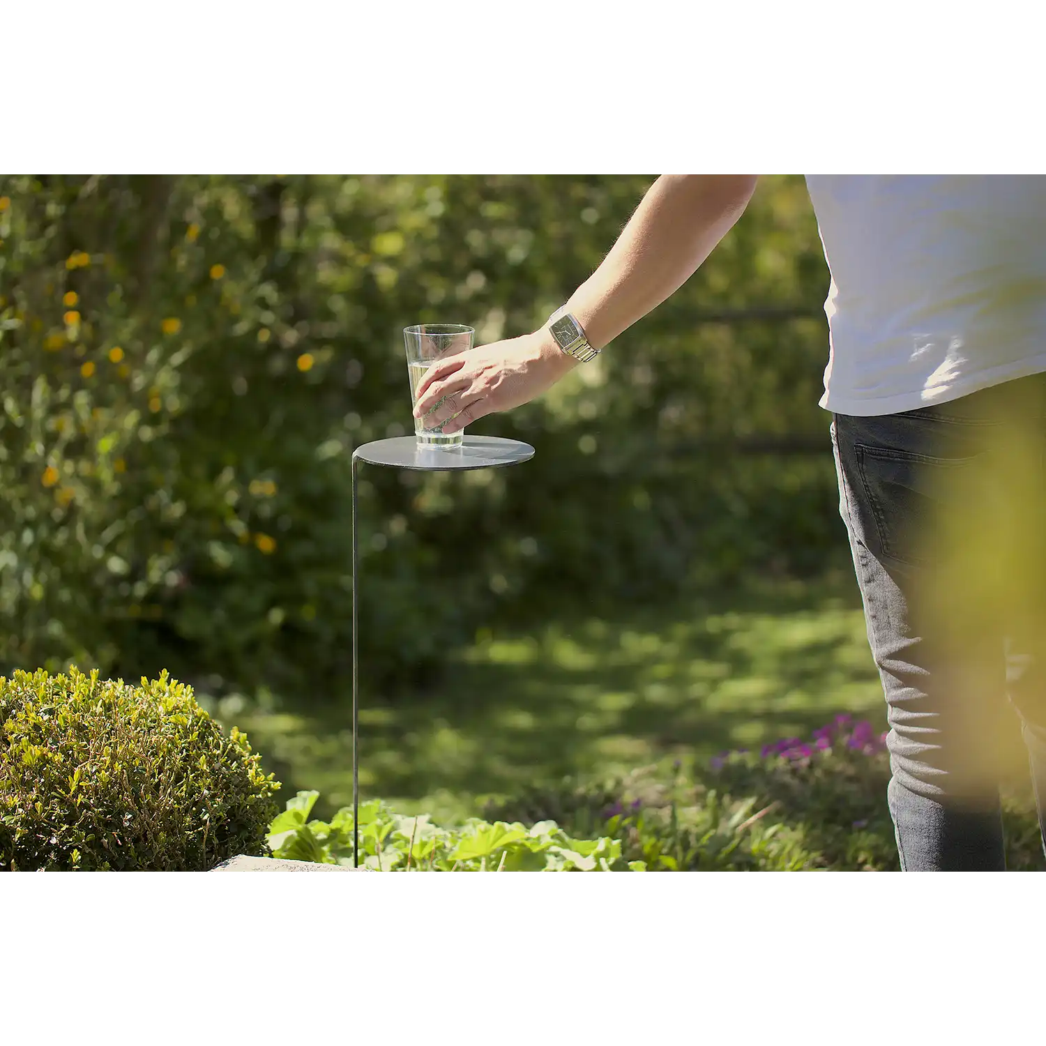 Eine Person im weißen Hemd stellt ein Glas Wasser auf das MONO Gartentischchen aus geöltem Stahl in einem von grünen Pflanzen und Büschen umgebenen Garten.