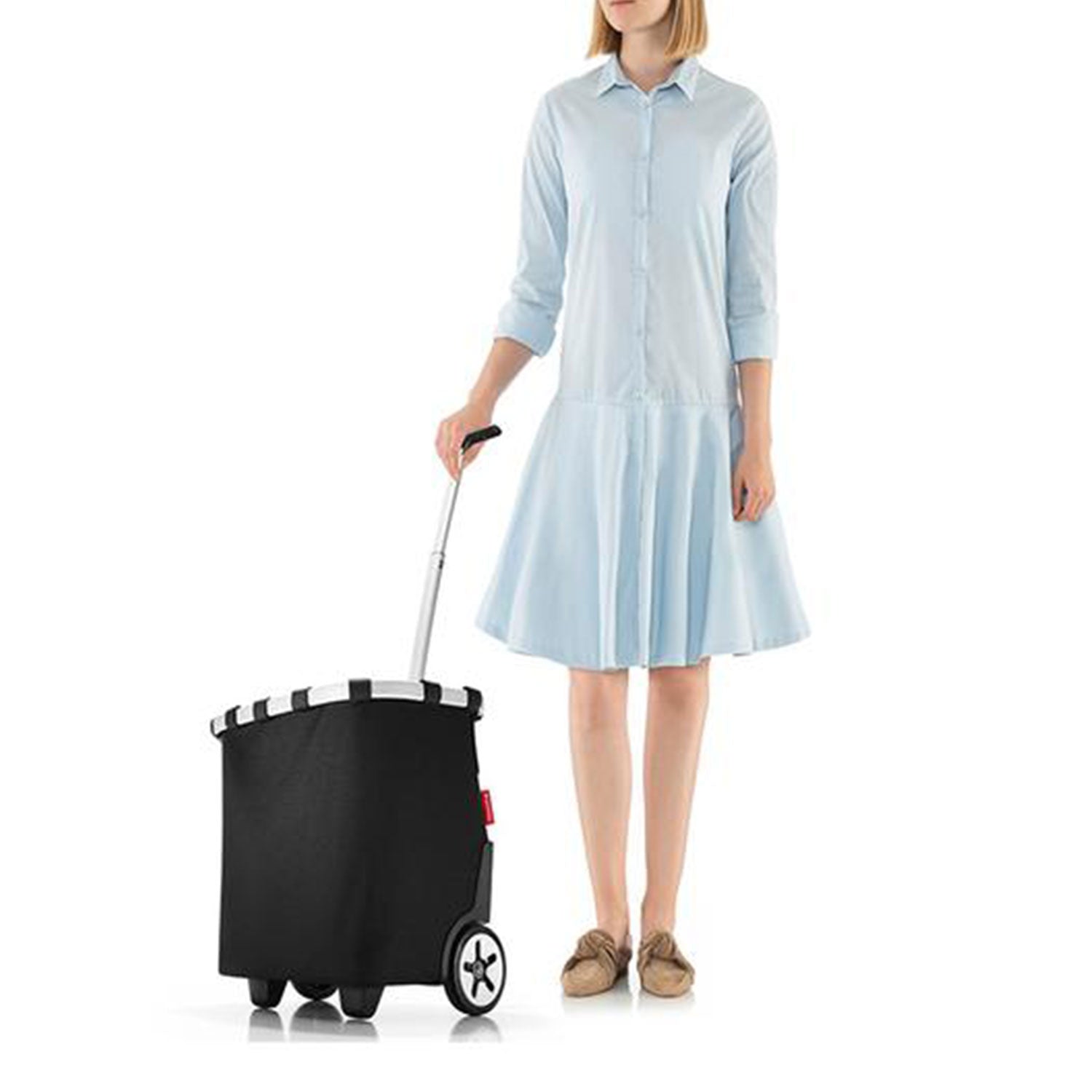 Eine Person in hellblauem Kleid und braunen Schuhen hält elegant den zweirädrigen Reisenthel Carrycruiser® – schwarz mit silbernem Teleskopgriff und gestreifter Felge. Vor dem weißen Hintergrund wirkt er leicht.