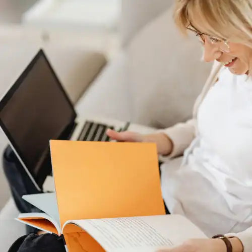 Eine lächelnde Frau liest ein Buch mit einem Laptop auf dem Schoß.