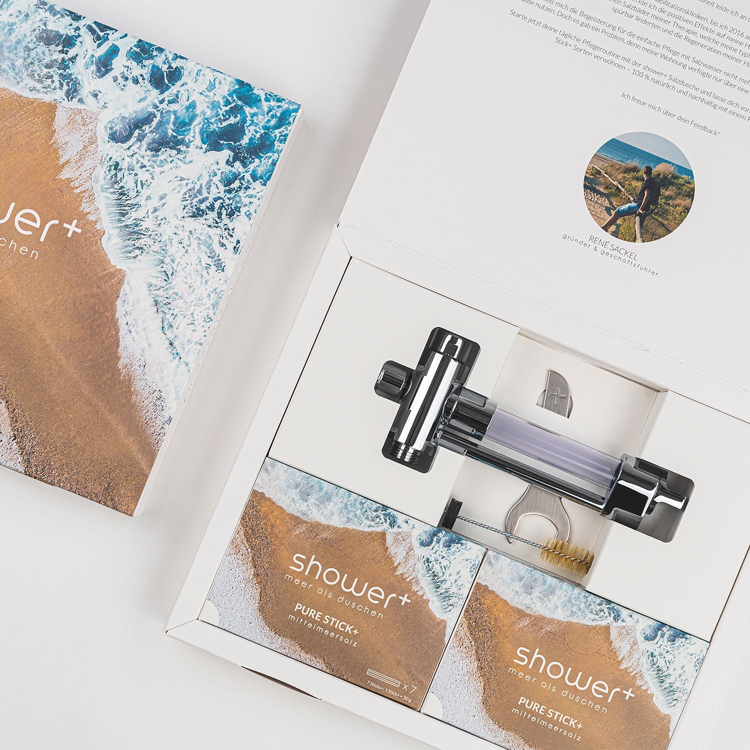 Offene Verpackung für „Deine Salzdusche für Zuhause“, ein Duschfilter-Set für empfindliche Haut, mit zwei Kartuschen, einem Metallgehäuse und Zubehör. Die Verpackung zeigt ein Strandmotiv mit der Aufschrift „shower+“ auf Deckel und Abdeckung.