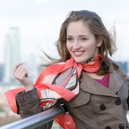 Lächelnde Frau in Mantel und buntem Schal, die im Freien steht, mit der Skyline der Stadt im Hintergrund.