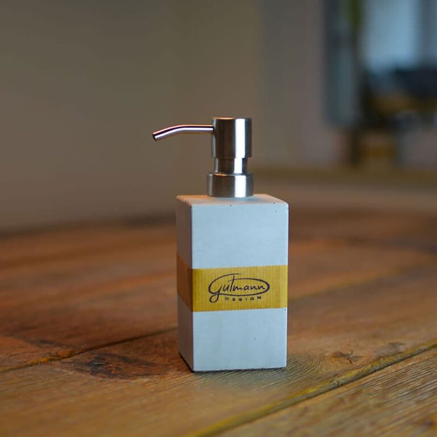 Ein Seifenspender aus Beton mit silberner Pumpe steht auf einer Holzoberfläche und ist mit einem gelben Etikett mit schwarzer Schrift verziert. Er verleiht der Badezimmer-Accessoire-Kollektion Eleganz, während der Hintergrund sanft verschwommen bleibt.