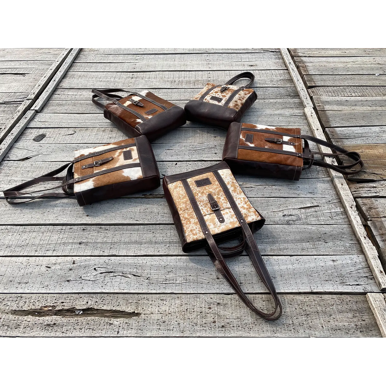 Fünf Shopper HALLODRI-Ledertaschen von Alpenleder mit einzigartigen braunen und gemusterten Unikat-Kuhfell-Einsätzen und dunklen Griffen sind elegant sternförmig auf verwitterten Holzbrettern angeordnet, wobei die Griffe nach außen zeigen.