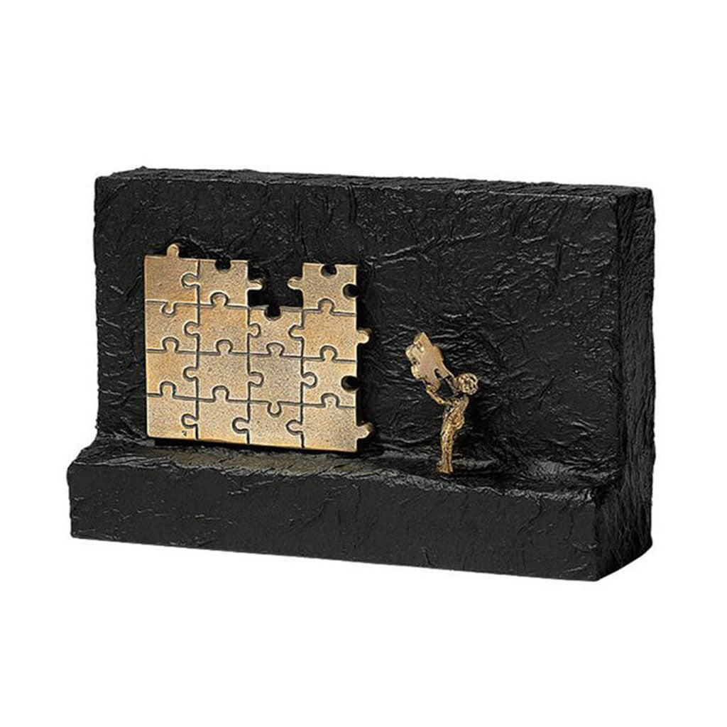 Die Skulptur „Seinen Teil beitragen“ ist ein strukturierter schwarzer Block mit einer bronzenen Puzzleteil-Skulptur, die auf einer Seite teilweise zusammengesetzt ist. Sie zeigt eine kleine Bronzefigur, die das letzte Teil an seinen Platz schiebt und so Vollendung und Beständigkeit symbolisiert.