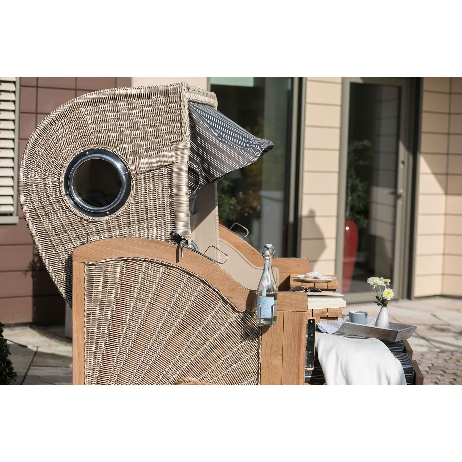 Ein Strandkorb DÜNE, grau mit Nadelstreifen, mit einem Baldachin aus PE-Geflecht steht im Freien. Ein Beistelltisch daneben bietet Platz für ein Glas Wasser, eine Zeitschrift und eine gefaltete Decke. Eine kleine Vase mit Blumen verleiht dem Ganzen Charme, während im Hintergrund friedlich ein beiges Gebäude thront.