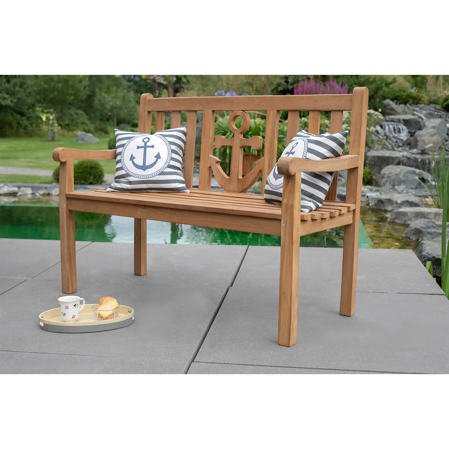 Die Teak-Gartenbank Anker im Anker-Design wertet Ihren Außenbereich auf, begleitet von zwei gestreiften Anker-Kissen. Ein rundes Tablett bietet Platz für Tassen und Gebäck, während ein Garten und ein kleiner Teich die ruhige Atmosphäre vervollständigen.
