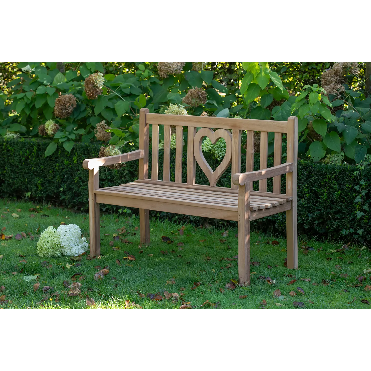 Die Teak-Gartenbank Herz mit herzförmiger Rückenlehne steht auf Gras, umgeben von Hortensien und grünen Büschen. Neben dieser Sitzgelegenheit im Freien steht ein Strauß weißer Blumen auf dem Boden.