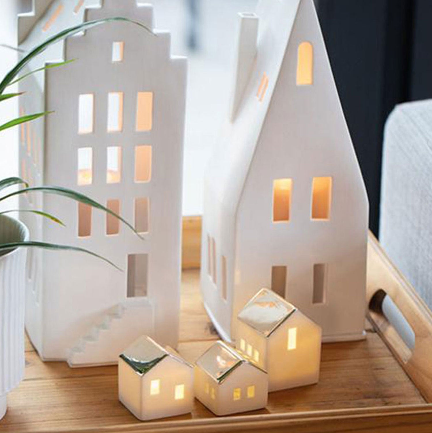 Auf einem Holztablett stehen Teelichthaus-Kerzenhalter aus unglasiertem Porzellan, darunter das Große Lichthaus „Giebeldach“, in dessen Inneren warmes Licht leuchtet. Daneben sorgt eine kleine Topfpflanze für eine behagliche Atmosphäre.