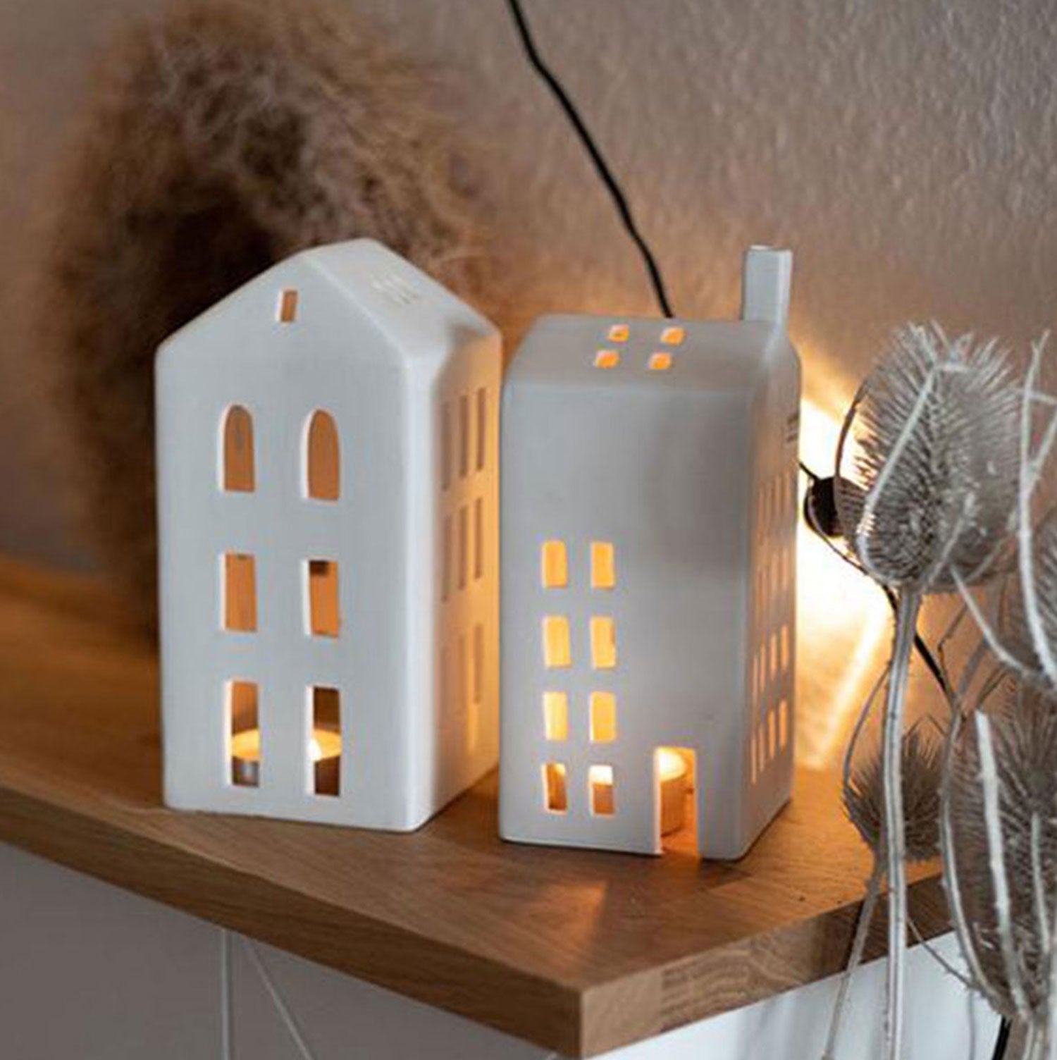 Das Lichthaus „Mittel, Kleine Fenster“ sind Teelichthalter aus Keramik, die an urige Häuser erinnern. Angezündet strahlen sie ein warmes Licht durch ihre Fenster auf einem Holzregal und bilden zusammen mit dekorativen Trockenpflanzen eine charmante Weihnachtsdekoration.