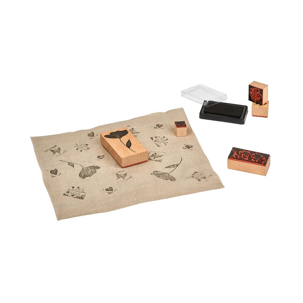 Mit einem Textilstempel-Set können Sie ein DIY-Projekt mit schwarzen Blatt- und Blumenmustern auf Stoff erstellen. Dabei werden verschiedene Stempeldesigns ordentlich angeordnet und ein Stempelkissen liegt in der Nähe.