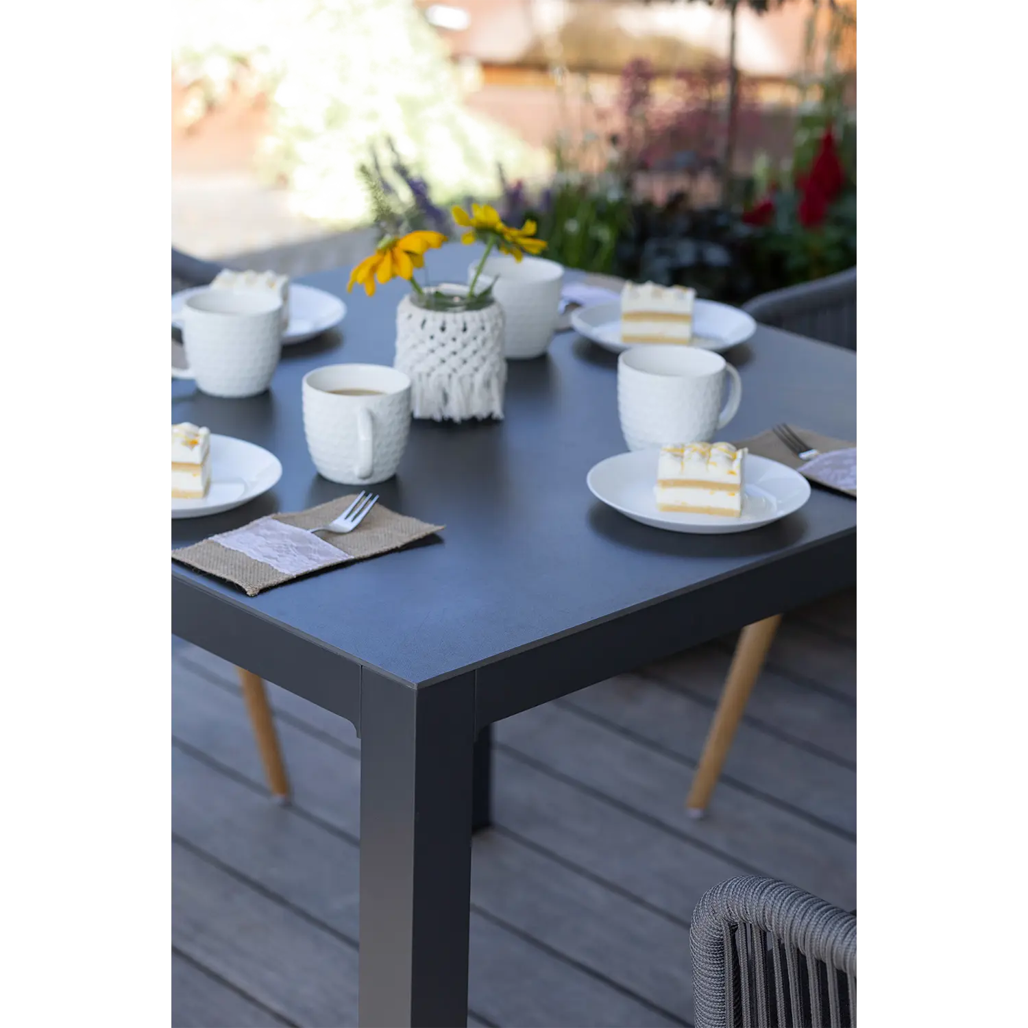 Ein Tisch im Freien auf einem robusten, wetterfesten Tisch Tarifa bietet vier Stücke Schichtkuchen auf weißen Tellern mit passenden Tassen. Eine kleine Vase mit leuchtend gelben Blumen ziert die Mitte, umgeben von üppigen Pflanzen auf der Holzterrasse.